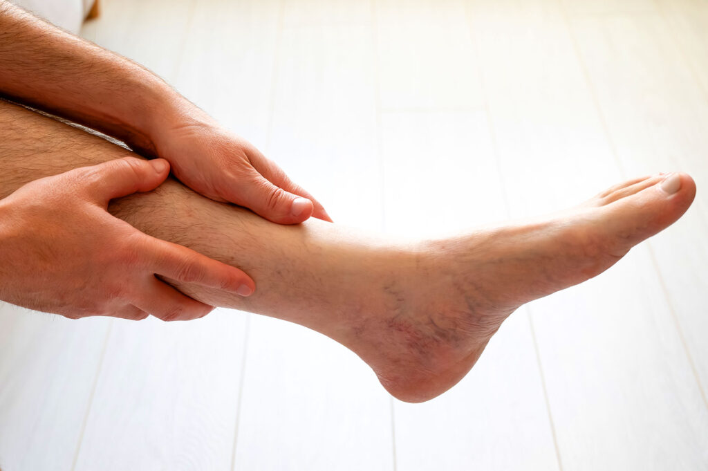 A close-up of a person's hands massaging their lower leg and ankle area over a light wooden floor.