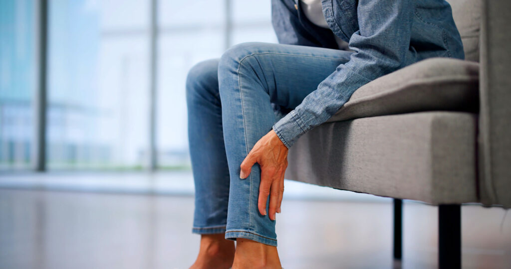 A person sitting on a sofa and holding their lower leg while experiencing discomfort or pain.